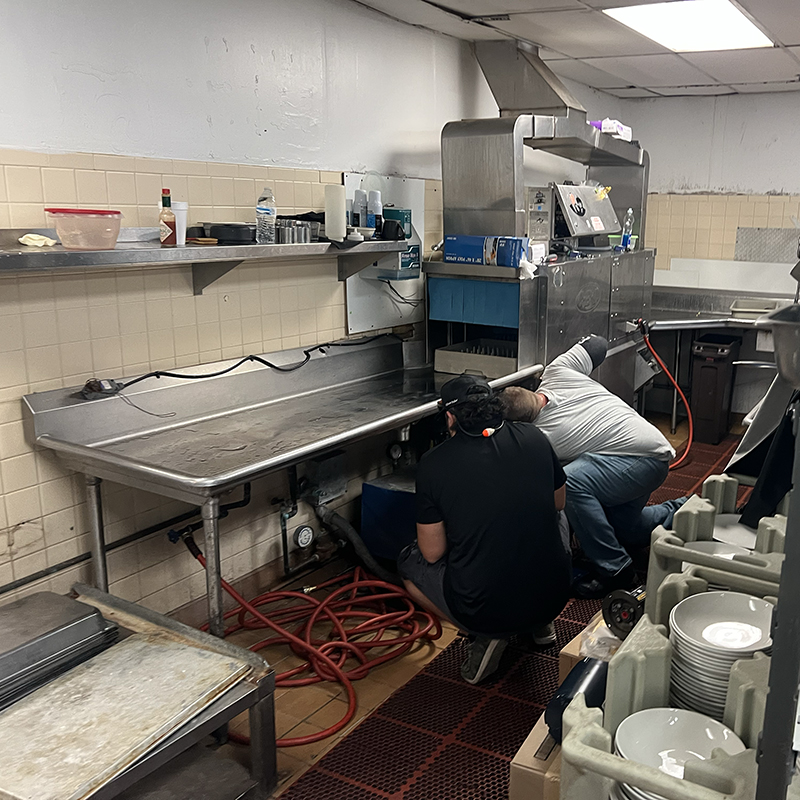 Two technicians repairing a commercial kitchen appliance in a restaurant setting, surrounded by stainless steel surfaces, cleaning supplies, and dishware.